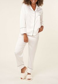 Ensemble de pyjama en satin blanc avec des passepoils noirs, comprenant des manches longues, une fermeture à boutons et un pantalon droit. Associé à des chaussons moelleux roses.