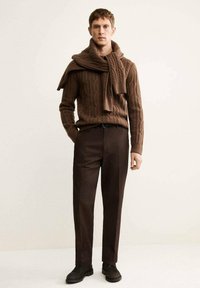 Maglione marrone a maglia con un motivo a treccia, abbinato a pantaloni marroni scuri. Il maglione ha una sciarpa drappeggiata in modo informale sulle spalle.