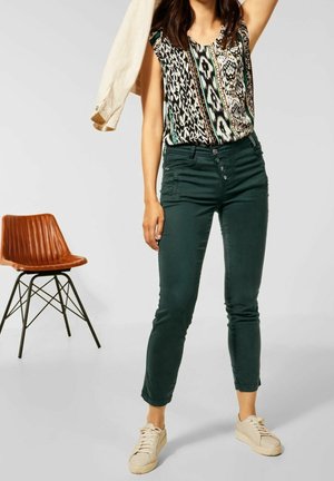 Donna con top senza maniche a fantasia, pantaloni aderenti verde scuro con bottoni, sneakers beige, che tiene una giacca beige sulla spalla.