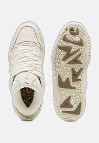 Krämfärgade sportskor med texturerad tyg, platta snören, prägling av varumärket och mönstrad gummisula med cirkulära mönster och detaljer.