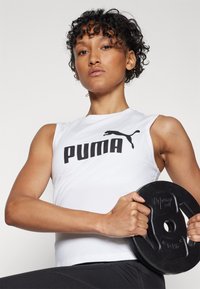 Hvit ermeløs treningstopp med svart "PUMA"-logo, kombinert med svarte treningsshorts. Personen holder en svart vektskive.