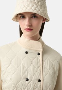 Pikowana kamizelka w kolorze kremowym z prążkowanym kołnierzem i dwoma czarnymi guzikami. Dopasowany pikowany kapelusz typu bucket. Gładka faktura i faliste detale wzoru.