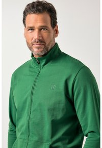 Grüne Reißverschlussjacke mit hohem Kragen, aus weichem Stoff. Verfügt über ein geprägtes Logo auf der Brust und eine glatte Textur.