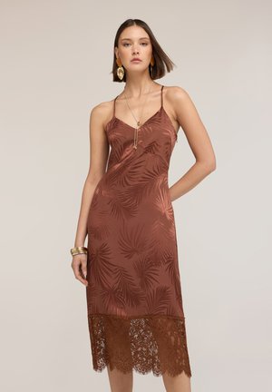 Mujer con vestido marrón de satén sin mangas con estampado de hojas y dobladillo de encaje, complementado con joyas doradas y cabello corto.