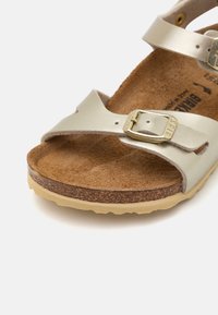 Sandal de couro dourado claro com duas tiras ajustáveis e uma fivela de metal dourado. Palmilha de cortiça texturizada e sola de borracha leve.