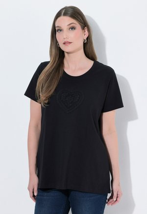 Frau mit langen braunen Haaren, die ein schwarzes T-Shirt mit einem strukturierten Herzdesign und blaue Jeans trägt, steht vor einem weißen Hintergrund.