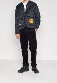 Veste matelassée noire avec des boutons-pression, ornée d'un patch sourire jaune et de détails textuels. Portée sur un pantalon côtelé sombre et des chaussures.