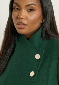 Vihreä villakangastakki, jossa on korkea kaulus, kaksirivinen malli ja kaksi pyöreää kultanappia. Silkkinen pinta ja muotoiltu siluetti.