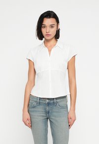 Λευκό πουκάμισο με κοντό μανίκι, κούμπωμα με κουμπιά μπροστά και κάθετη ριγέ υφή. Εφαρμοστή σχεδίαση με γιακά, συνδυασμένο με ανοιχτόχρωμο μπλε τζιν.