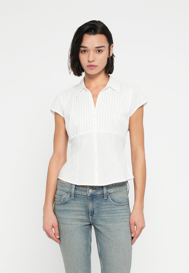 Λευκό πουκάμισο με κοντό μανίκι, κούμπωμα με κουμπιά μπροστά και κάθετη ριγέ υφή. Εφαρμοστή σχεδίαση με γιακά, συνδυασμένο με ανοιχτόχρωμο μπλε τζιν.