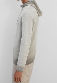 Sudadera de punto gris claro con una textura suave, que presenta una capucha en dos tonos y puños acanalados. El diseño incluye una cremallera frontal y bolsillos.