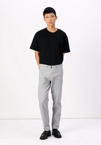 Camiseta negra de algodón con cuello redondo, combinada con pantalones gris claro. Llevado con zapatos negros, el modelo está de pie contra un fondo blanco liso.