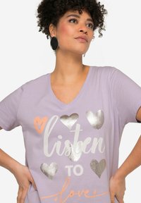 Camiseta de manga corta en color lavanda con escote en V, que presenta gráficos de corazones en plata y melocotón, y el texto "escucha al amor". Textura de algodón suave.