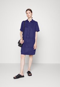Vestido de mangas curtas e colarinho em azul profundo, feito de um tecido leve, com fecho de botão na frente e dois bolsos frontais. Combinado com sandálias pretas de deslizar.