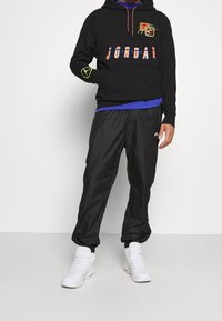 Persona que lleva una sudadera negra de Jordan con acentos neón, pantalones de chándal negros de Nike y zapatillas altas blancas de pie contra un fondo blanco.