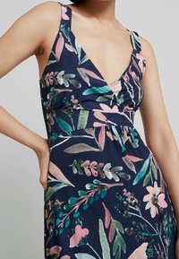 Sukienka w kwiatowy print z granatowym tłem, głębokim dekoltem w kształcie litery V oraz cienkimi ramiączkami. Wzór zawiera różowe i zielone liście oraz kwiaty.