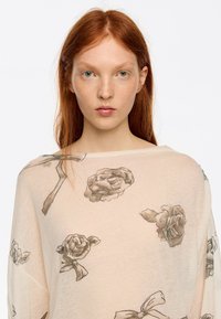 Lekki, beżowy sweter z miękkiego materiału, ozdobiony kwiatowym wzorem z szarymi różami i kokardkami. Luźny krój, szeroki dekolt i długie rękawy.