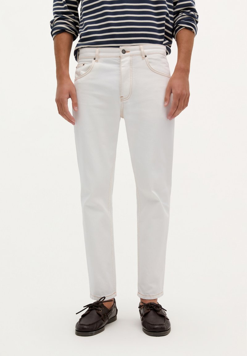 Pantalones de denim blancos con costuras naranjas, ajuste slim, diseño de cinco bolsillos, combinados con una camisa de manga larga a rayas en blanco y negro y zapatos marrones.