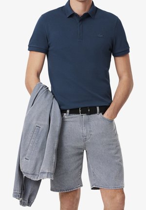 Homme portant un polo bleu marine et un short en denim gris clair tenant une veste gris clair assortie dans une main.