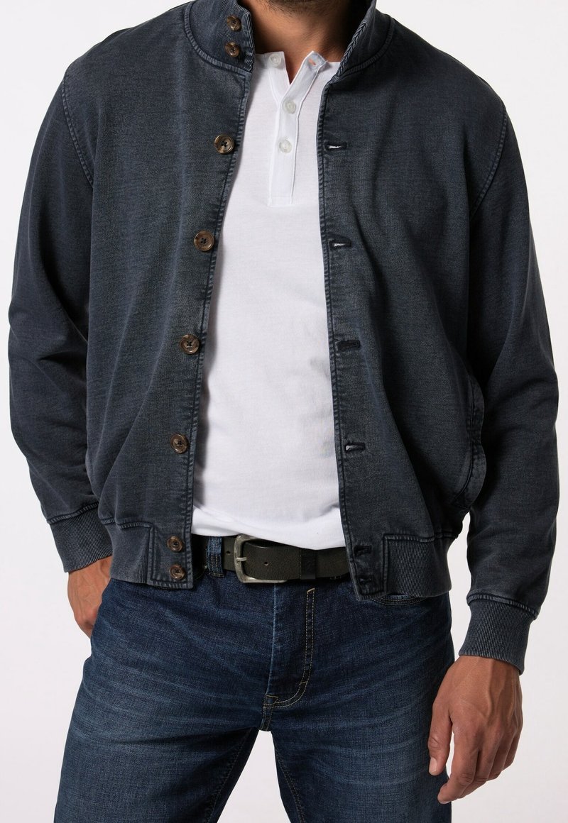 Homme portant une veste boutonnée bleu foncé sur une chemise henley blanche, jean bleu et ceinture noire, mains détendues le long du corps, sans détails en arrière-plan.