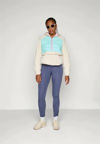 Lett og cropped hettegenser i krem og turkis med halv glidelås, sammen med mørk lilla leggings og beige joggesko. Stilige solbriller inkludert.