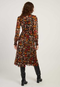 Vrouw met krullend haar, gekleed in een lang mouw, kalflengte jurk met oran jerode en beige bloemen- en dierenpatronen, gecombineerd met zwarte haklaarzen.