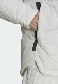 Main glissant dans la poche latérale d'une veste blanche matelassée avec une finition noire à l'ouverture de la poche, montrant la manche et la zone de la taille.