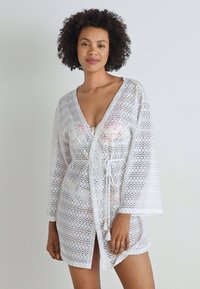 Cárdigan blanco de ganchillo con escote en V, mangas largas y cintura ajustable. Presenta patrones florales en su interior, acentuando su textura ligera.