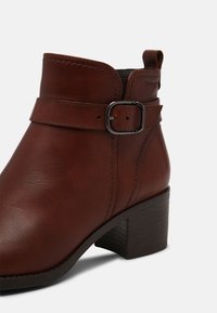 Hnědá kožená kotníková bota s kulatou špičkou, ozdobnou přezkou a vrstveným podpatkem. Obsahuje jemné švy a hladkou texturu.