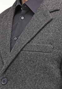 Muž ve tmavě šedém tvídovém saku přes černou košilí s knoflíky, ukazující detaily klopy saka, límce a náprsní kapsy.