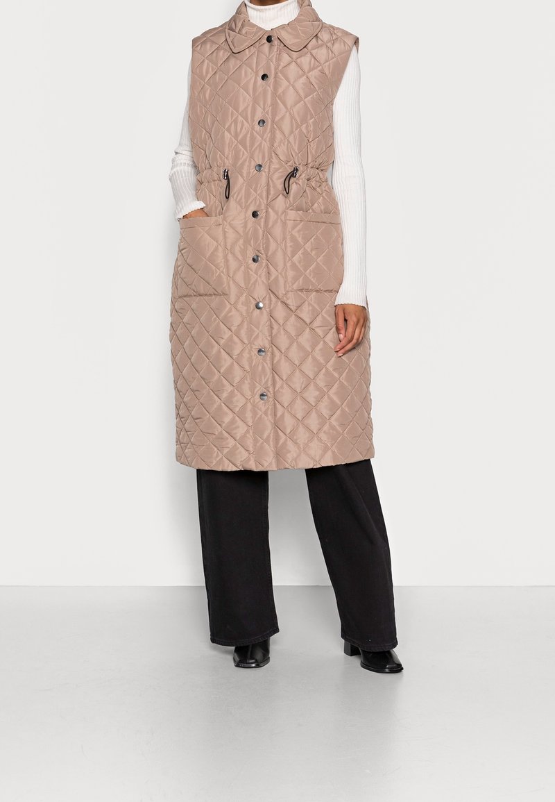 Gilet sans manches en beige matelassé avec un col, deux poches avant et une taille ajustable par cordon, associé à un pantalon large noir.