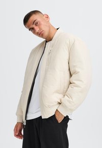 Puffer jacket in light cream with ribbed cuffs and collar, featuring a front zipper and a side pocket for storage.