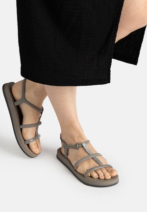 Füße, die braune flache Sandalen mit mehreren dünnen Riemen und einer Schnalle tragen, gezeigt mit einem schwarzen strukturierten Rock.