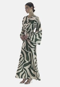Robe maxi en tissu vert et crème avec un design fluide, dotée de longues manches évasées et de motifs abstraits. Associée à des talons avec lanières.