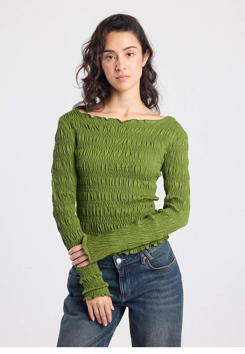 Green smocked long-sleeve top with an off-the-shoulder neckline and textured detailing. Paired with high-waisted blue denim jeans.