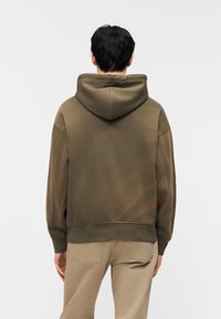 Άτομο που φοράει φούτερ με κουκούλα σε ελιά πράσινο χρώμα και μπεζ παντελόνι, στέκεται με την πλάτη προς το μέρος μας μπροστά σε απλό ανοιχτό φόντο.