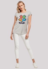 Grå bomulls t-skjorte med et fargerikt Hogwarts-våpenskjolddesign og ulike mønstre, kombinert med hvite skinny jeans og hvite sneakers.