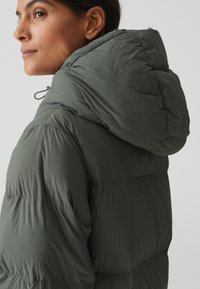Pufferjack in donkergroen met een lichtgewicht, gewatteerd ontwerp. Voorzien van een capuchon en elastische trekkoorden voor aanpassing. Zachte, iets gekreukt textuur.