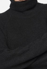Pull à col roulé en maille épaisse noire avec un motif côtelé texturé, couvrant le cou et les épaules, porté par une personne.