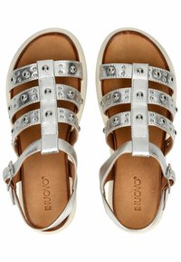 Stříbrné sandály z faux kůže mají několik širokých pásků se stříbrnými kulatými nýty, nízkou bílou podrážku a hnědý interiér s přezkou.