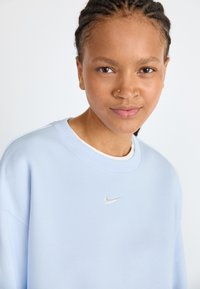 Světle modrá mikina vyrobená z měkké látky, s kulatým výstřihem a stříbrným logem Nike vyšitém na hrudi.