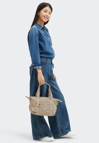 Mujer con camisa de denim y vaqueros de pierna ancha sosteniendo un bolso beige con llavero, sonriendo, de pie sobre un fondo blanco.
