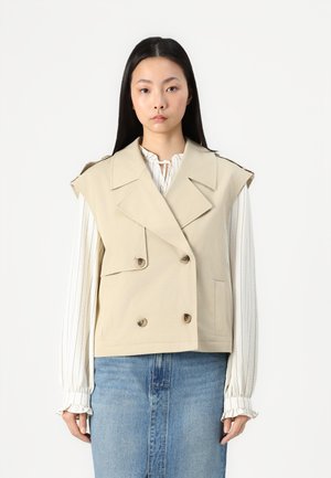 Beige doppelreiher Weste mit breiten Revers und Schulterpolstern, mit vorderen Taschen und braunen Knöpfen, getragen über einer gestreiften Bluse.