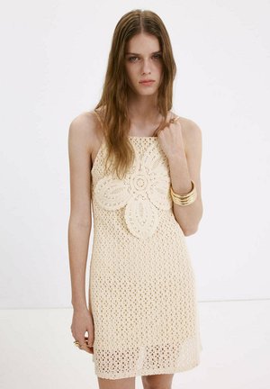 Mujer con cabello largo y castaño que lleva un vestido de crochet beige sin mangas con un gran patrón de flores y pulseras doradas en la muñeca.