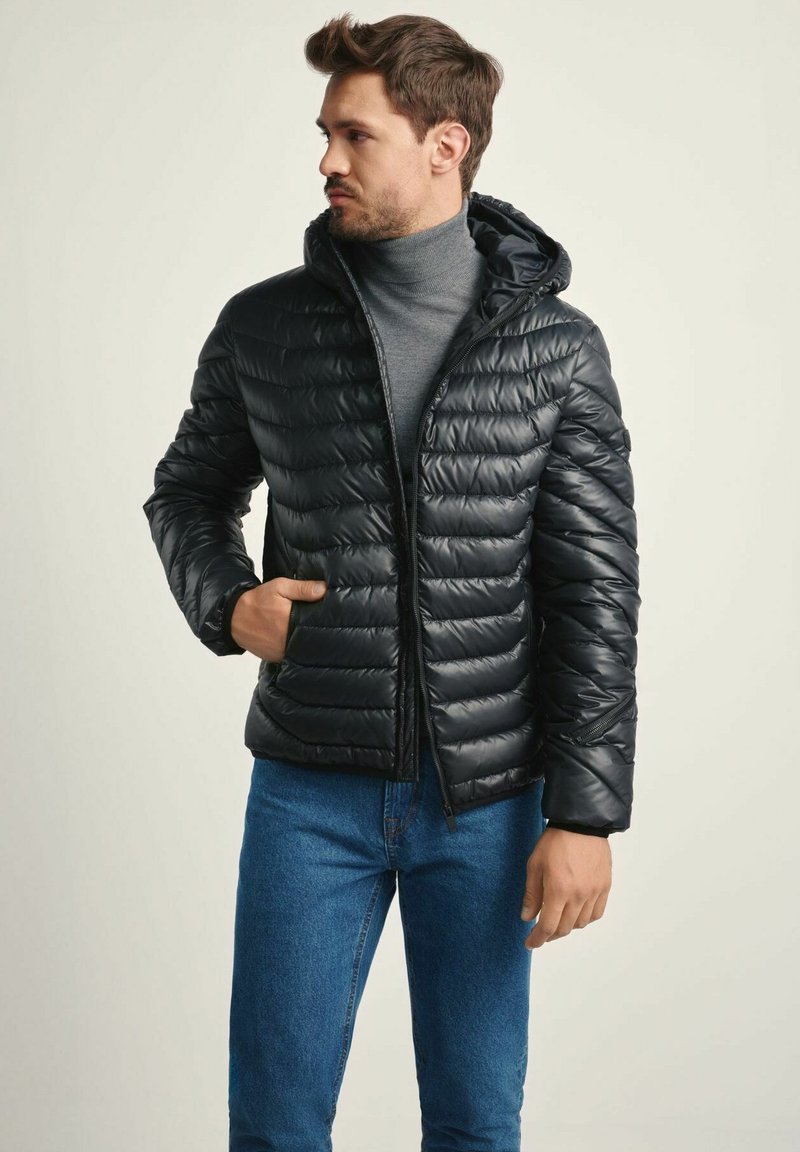 Czarna kurtka puchowa z kapturem, z pikowanym wzorem, błyszczącą fakturą i kieszeniami na zamek. Nosi się ją na szary golf i niebieskie dżinsy.