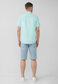 Licht turquoise shirt met korte mouwen en een relaxte pasvorm, gecombineerd met lichtblauwe denim shorts en witte sneakers, gezien van achteren.