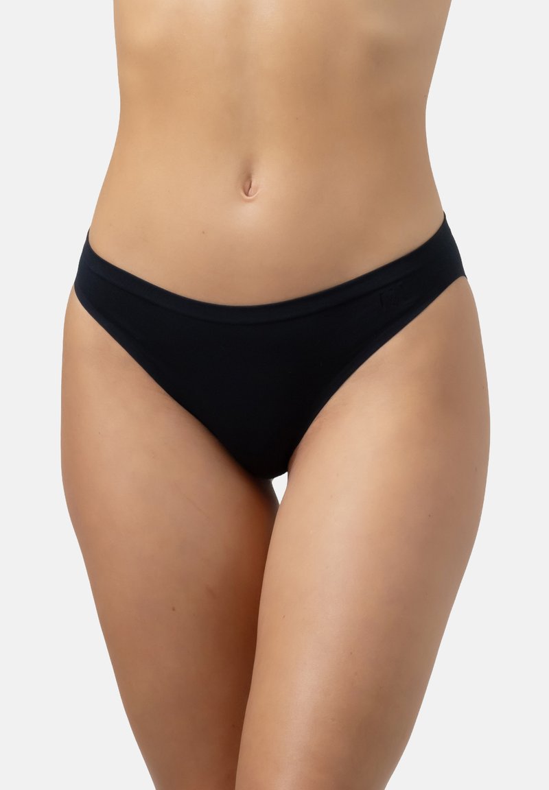 Vrouw in zwarte naadloze bikini-achtige onderbroek, die het midden van de buik en de bovenste dijen toont tegen een effen witte achtergrond.