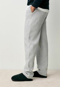 Pantaloni sportivi grigi chiari realizzati in morbido tessuto, con tasche laterali e una vestibilità ampia, abbinati a pantofole testurizzate verde scuro.