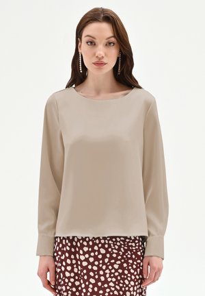 Donna con capelli castani lunghi indossa una blusa beige a maniche lunghe e una gonna color vinaccia con macchie chiare irregolari, in piedi davanti a uno sfondo bianco.