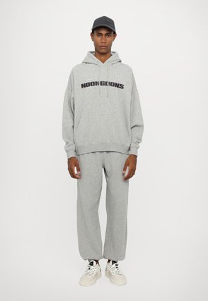 Hoodie gris avec le texte "NOONGOONS", associé à un pantalon de survêtement gris assorti. Porté avec des baskets blanches, à lacets ronds et une casquette de baseball.
